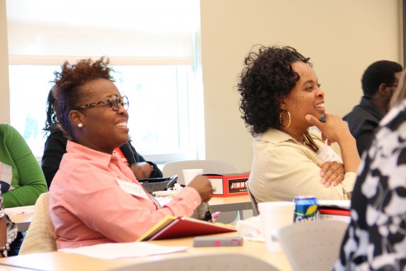 Two women listening to a speaker and smiling while sitting at a table with notebooks.